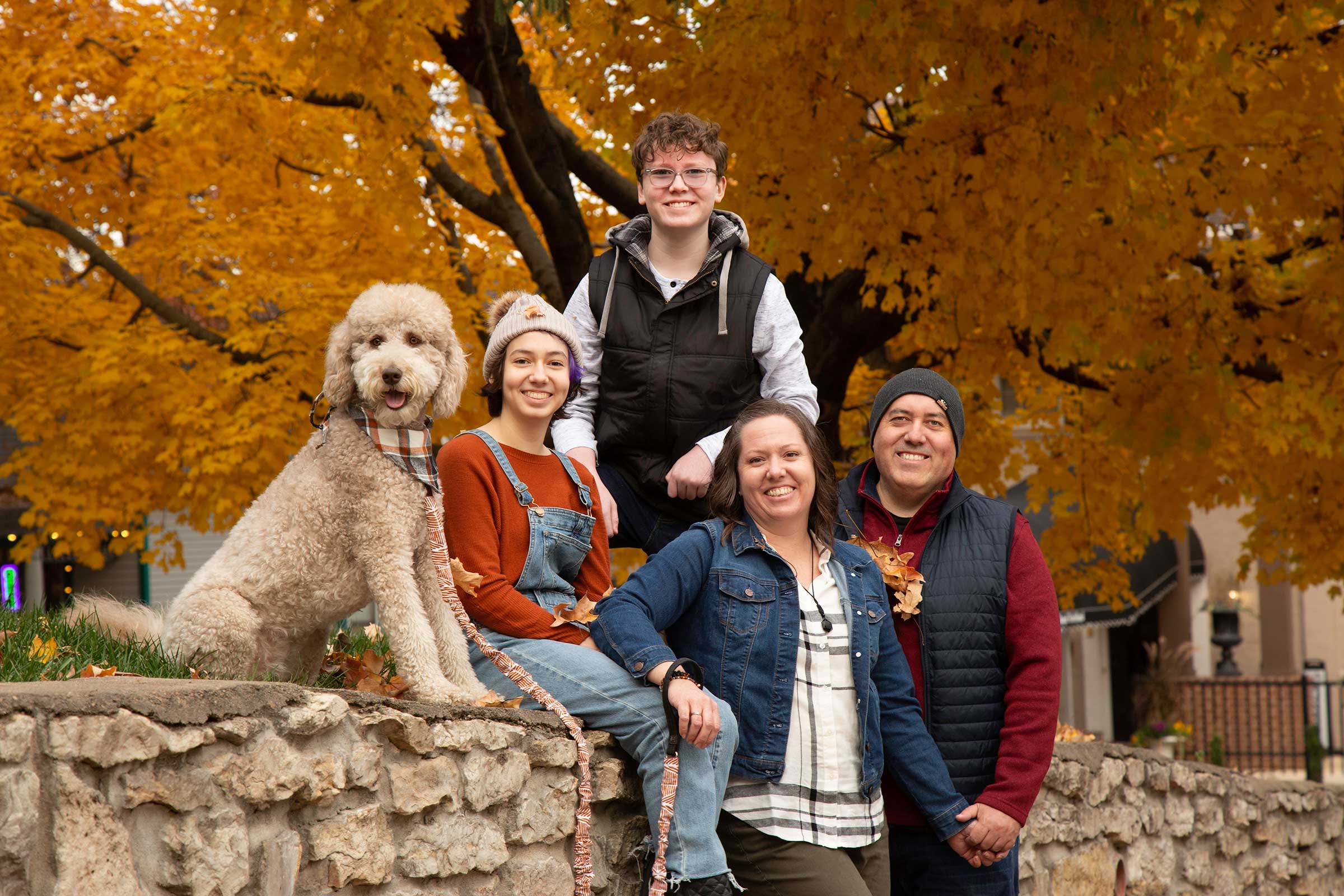 Family portrait in Parkville MO featuring parents, teenagers, and their dog posing outdoors in front of vibrant fall foliage
